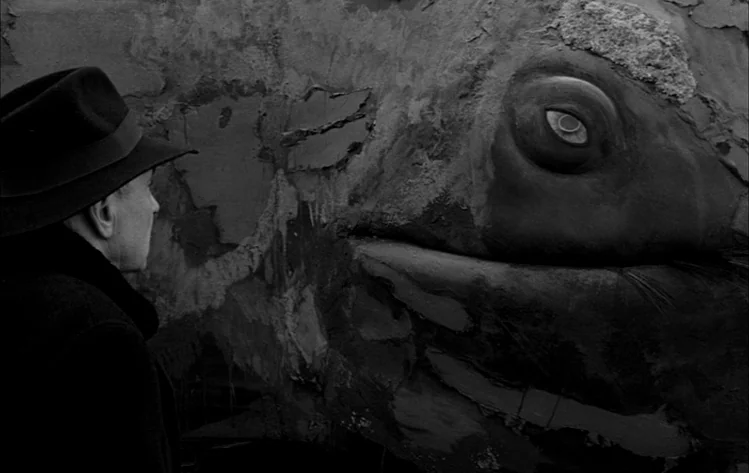 Werckmeister Harmonies (2000) movie still: medium — A man faces the massive, textured carcass of a whale resting on dark terrain. The whale's eye is…