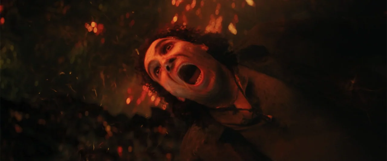 Black Adam (2022) movie still: closeup — A dark outdoor area filled with flying embers and smoke from nearby fires. A man with dirt on his face…
