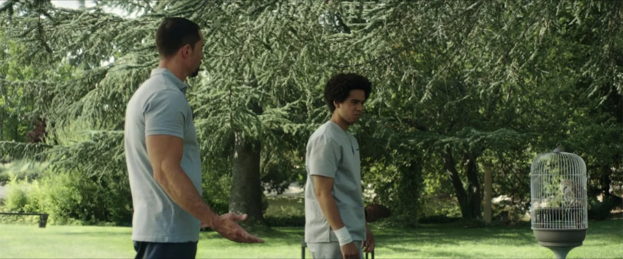 American Carnage (2022) movie still: medium — A man and a youth stand on a grassy lawn near large pine trees. The man gestures with his hand towards…