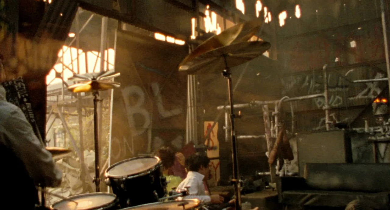 Burst City (1982) movie still: wide — A cluttered, industrial rehearsal space with exposed piping and graffiti-covered walls. Several musicians are…