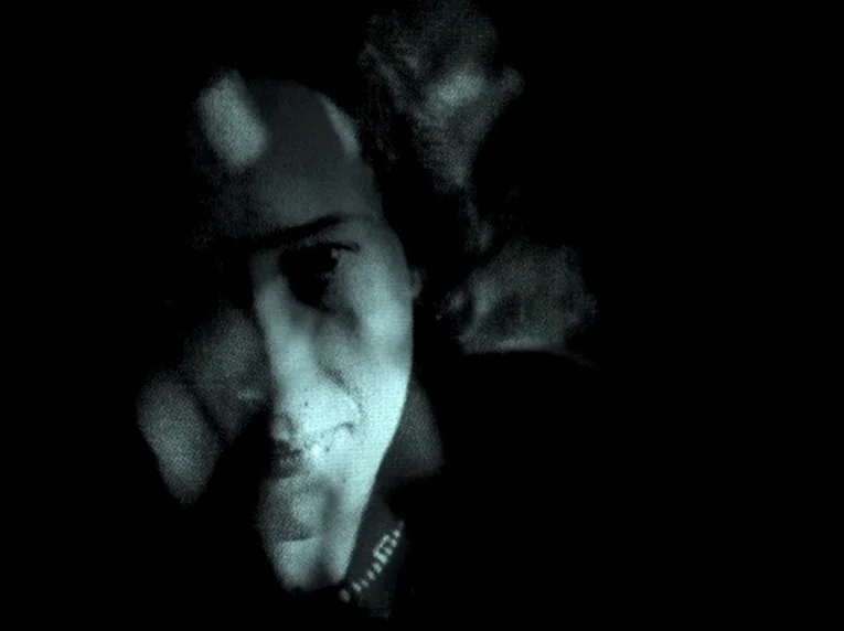 Notre Musique (2004) movie still: closeup — A dark, indistinct space surrounds a single individual illuminated by filtered light. A person's face…