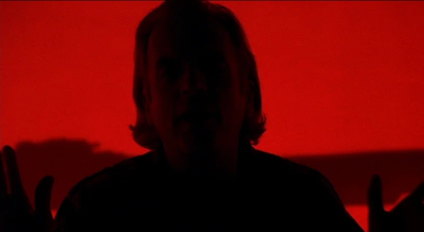 Gray’s Anatomy (1996) movie still: closeup — A man is centered in the frame against a flat, solid red background. The subject is backlit and appears…