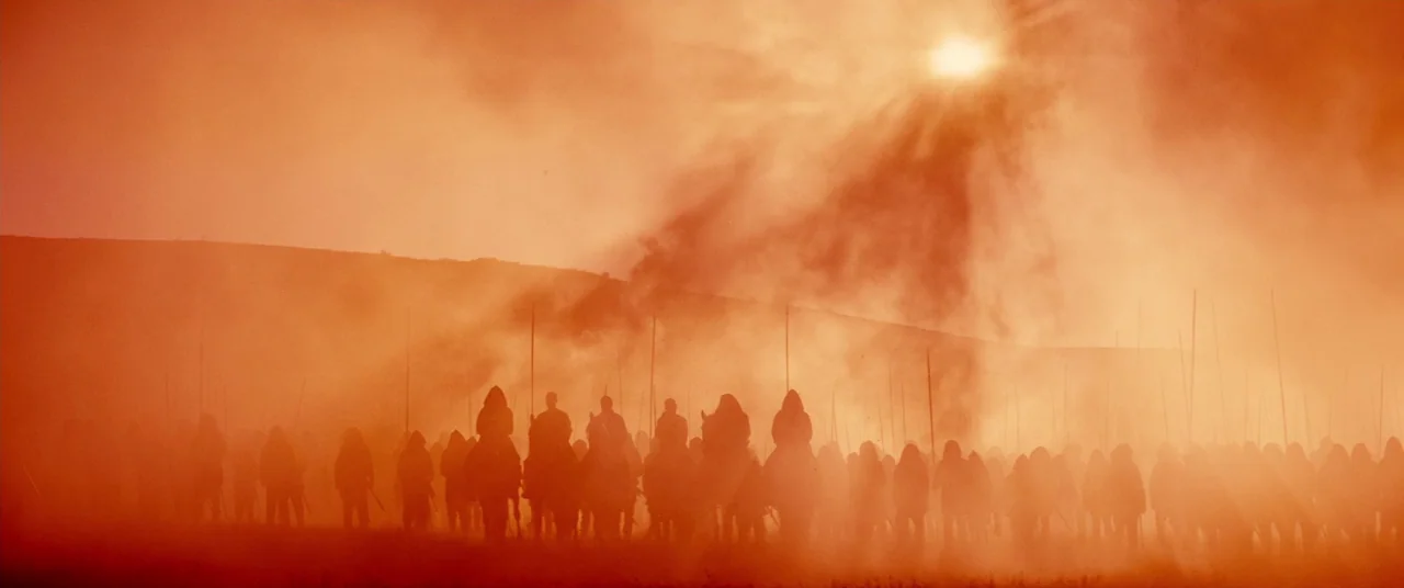 Macbeth (2015) movie still: wide — A vast crowd of silhouettes stands in a dusty, sun-bleached open landscape. The group holds long vertical spears…