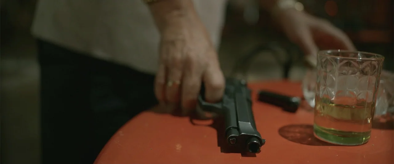 Divine Intervention (2026) movie still: closeup — A close view of a red table surface inside a dimly lit establishment. A hand rests beside a…