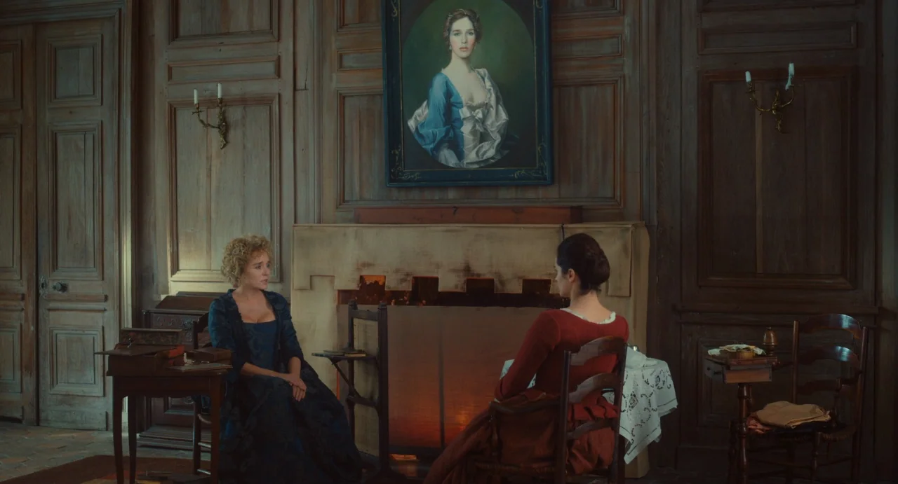 Portrait of a Lady on Fire movie still: wide — A wood-paneled room features a central fireplace topped by an oil painting of a woman. Two women are…