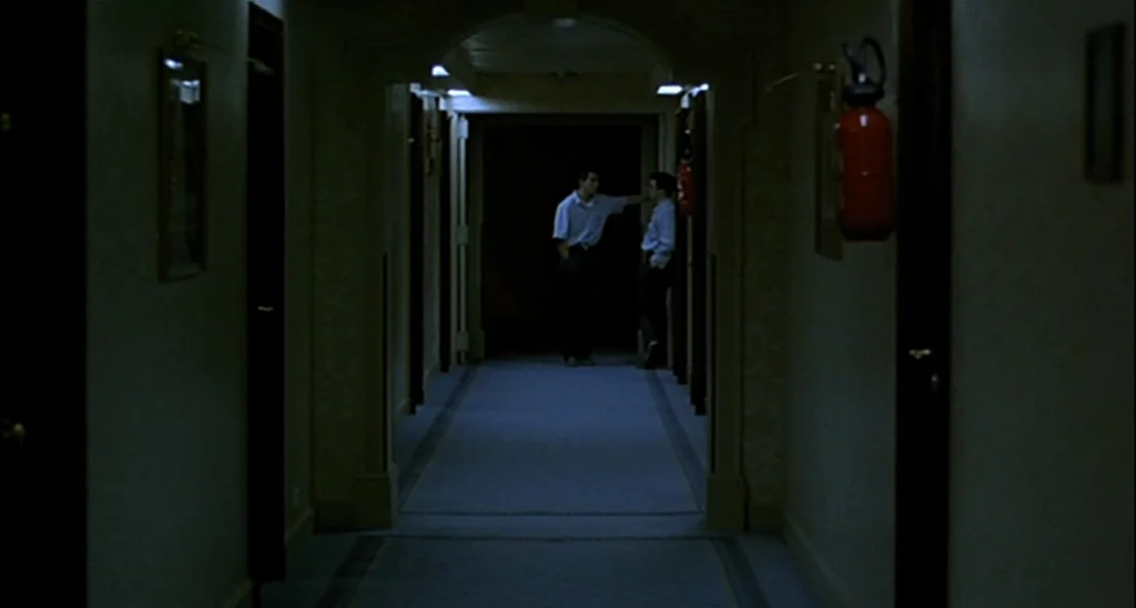 Trouble Every Day (2001) movie still: wide — A long, dimly lit hotel corridor features arched doorways and muted blue carpeted flooring. Two men…