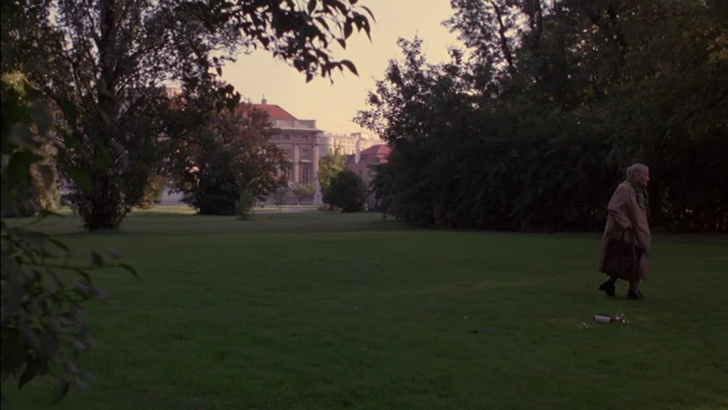 Before Sunrise (1995) movie still: wide — A spacious green lawn in a city park surrounded by large trees and buildings. An elderly woman in a trench…