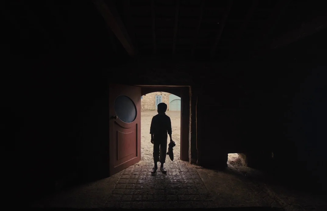 Gretel & Hansel (2020) movie still: wide — A dark stone barn has a large open doorway looking out onto a dirt courtyard. A small child stands in the…