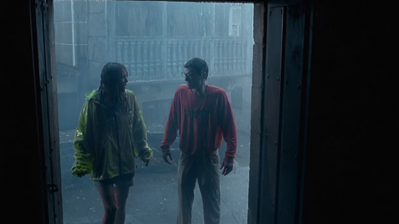 Dagon (2001) movie still: fullbody — Two people stand in a shadowed doorway overlooking a rainy cobblestone courtyard. Heavy rain falls on the…