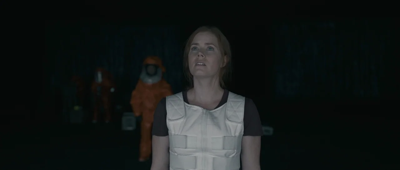 Arrival (2016) movie still: medium — A dark, cavernous space surrounds a woman in a white protective vest. Two figures wearing bright orange hazmat…