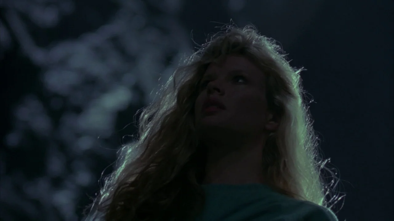 Batman (1989) movie still: closeup — The outdoor setting is dark with soft, diffused green light filtering through tree branches. A woman looks…