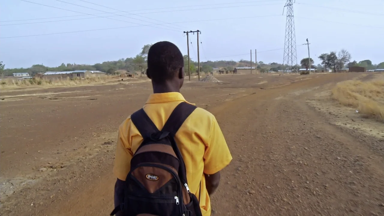 The Money Stone (2018) movie still: medium — A dry, open dirt road stretches across a rural landscape under an overcast sky. A young boy wearing a…