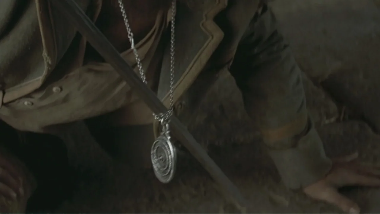 The Mask of Zorro (1998) movie still: closeup — A person in a drab brown coat kneels upon a dark, dusty dirt surface. A large circular metal pendant…