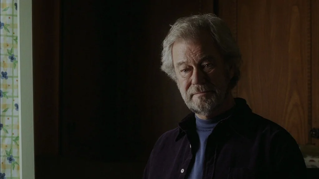 Away From Her (2006) movie still: medium — An interior space with wood cabinets and patterned floral wallpaper on the left. A middle-aged man stands…