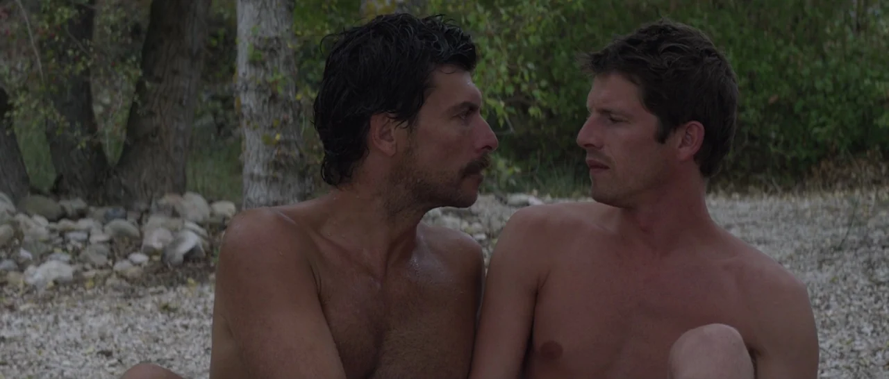 Stranger By The Lake (2013) movie still: medium — Two shirtless men sit on a pebbled beach surrounded by sparse vegetation and trees. The men are…