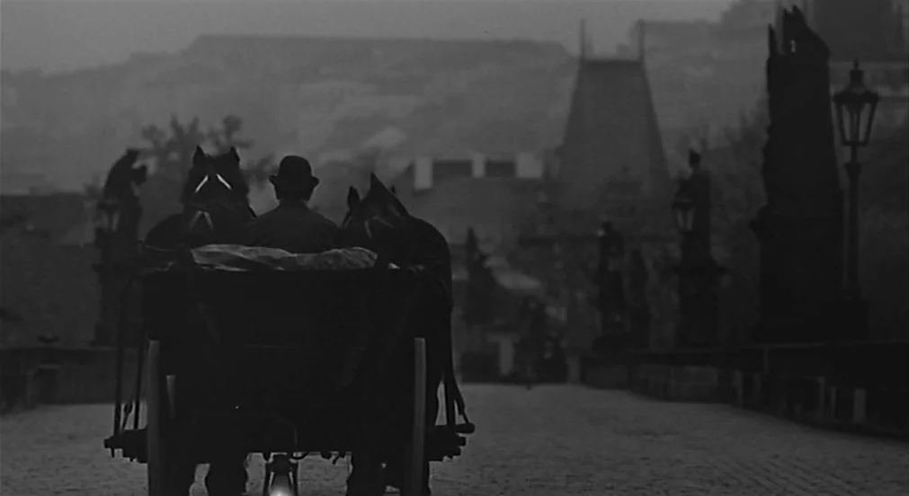 Kafka (1991) movie still: wide — An old stone bridge with statues lines the path under a grey morning sky. A man drives a horse-drawn carriage away…