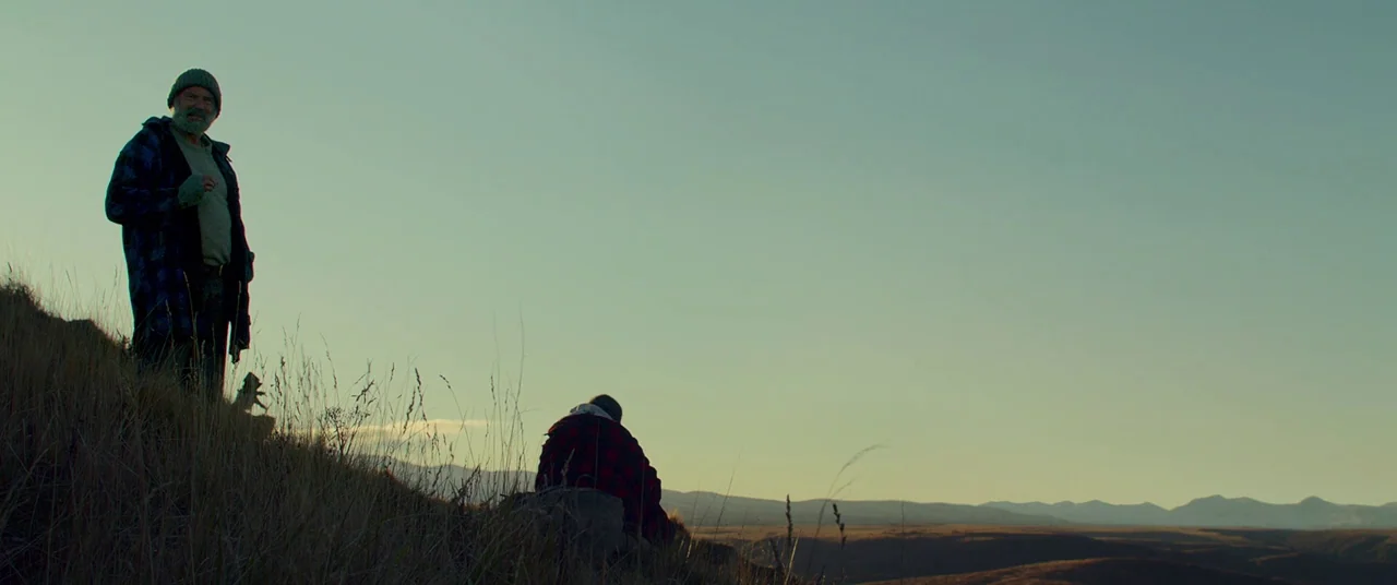 Hunt for the Wilderpeople (2016) movie still: wide — An open hillside covered in dry grasses overlooks a vast, hazy valley at dusk. A standing man…