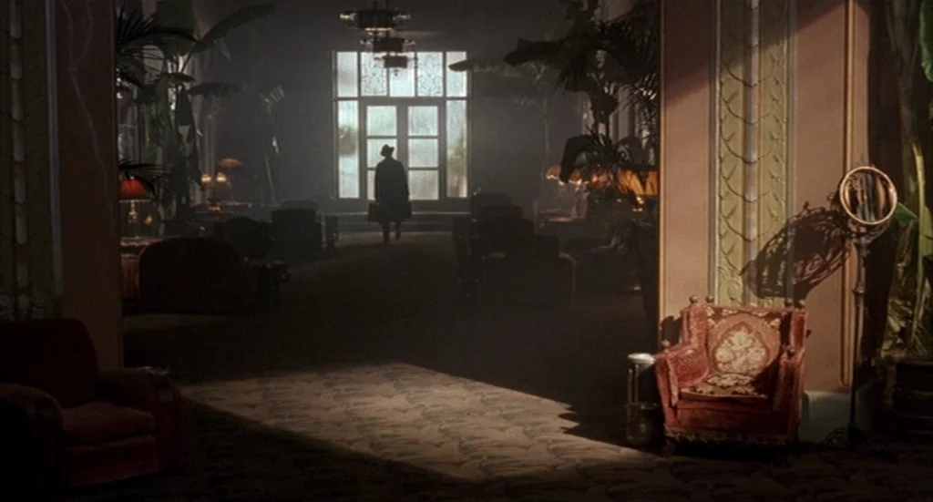 Barton Fink (1991) movie still: wide — A dimly lit, ornate interior lobby with decorative moldings and potted plants. A man in a long coat walks…