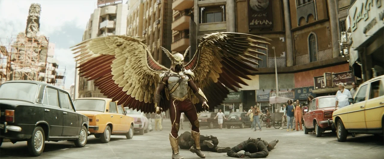Black Adam (2022) movie still: fullbody — A crowded urban square features vintage-style buildings, parked cars, and several bystanders in the…