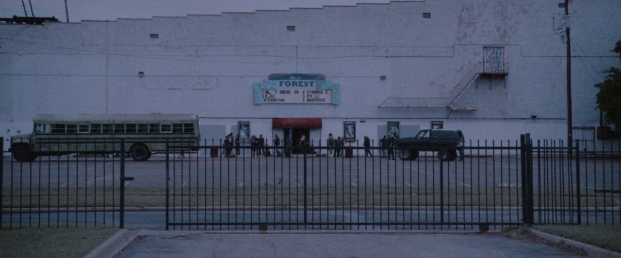 VFW movie still: establishing — A wide shot shows a large cinema facade at dusk behind a black iron fence. A group of people stands in a line…