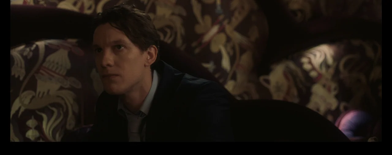 The Wrong Tenant (2022) movie still: medium — A dark, dimly lit room features a patterned sofa with floral upholstery designs. A young man in a dark…