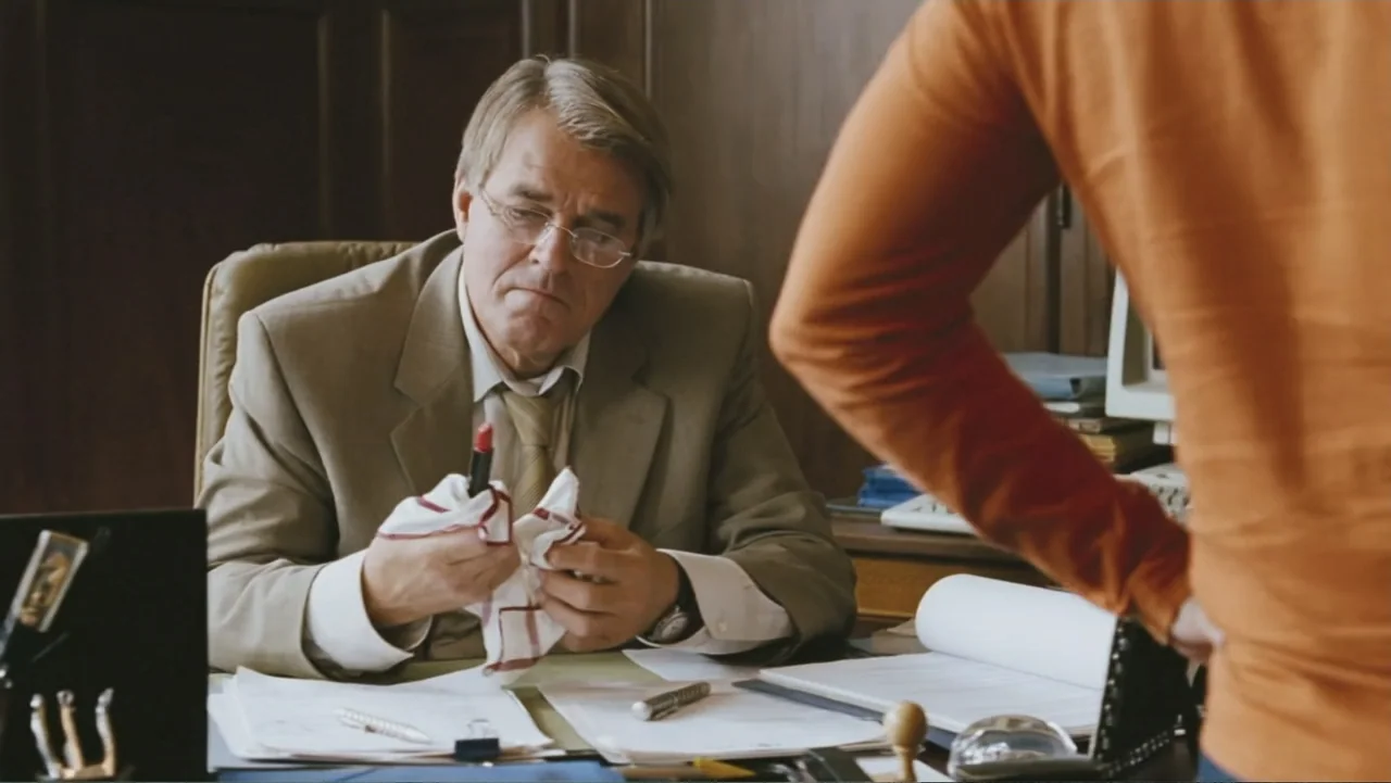 Deuce Bigalow: European Gigolo (2005) movie still: medium — A man sits at a cluttered wooden desk in an office with dark walls. The man examines a…