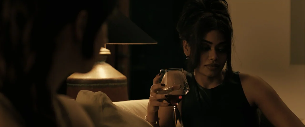 Companion (2025) movie still: medium — A dimly lit domestic interior features a woman sitting on a sofa with wine. The woman in focus holds a glass…