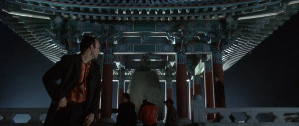 The Usual Suspects movie still: wide — A traditional Korean open-sided wooden structure with an ornate, multi-layered painted roof at night. A man…
