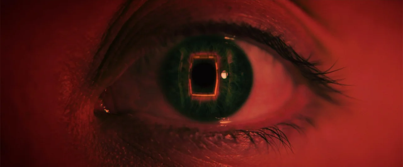 Nocturne (2020) movie still: closeup — A close-up view of a human eye captured in extreme red lighting conditions. The iris clearly reflects a…