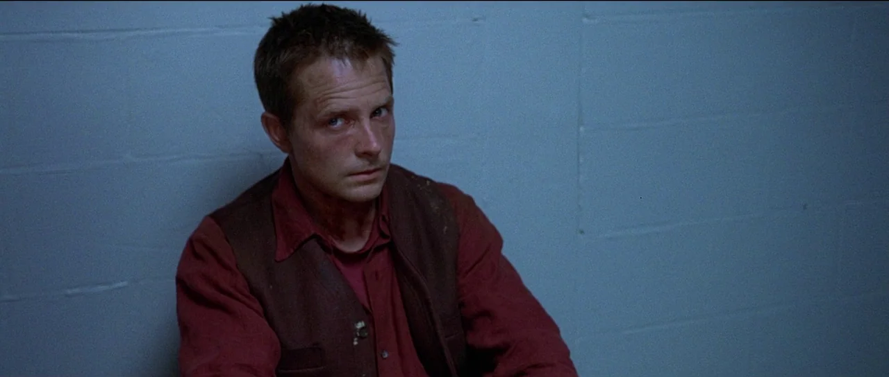 The Frighteners (1996) movie still: medium — A man sits against a pale blue painted concrete block wall. He wears a maroon shirt and dark vest while…