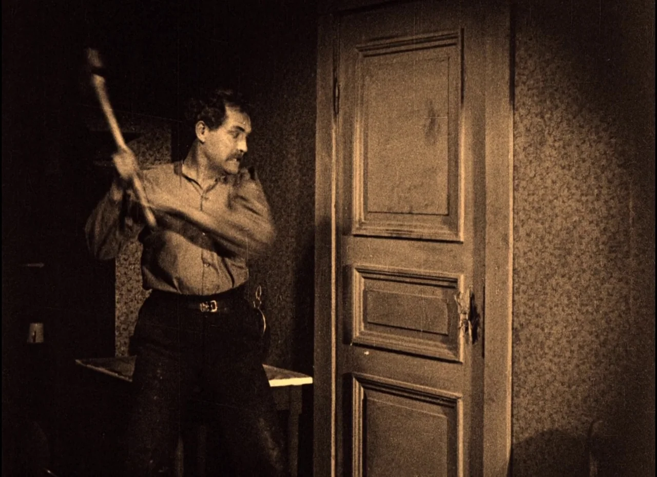 The Phantom Carriage (1921) movie still: medium — A dimly lit, cramped room features a heavy wooden door with rectangular molding details. A man…