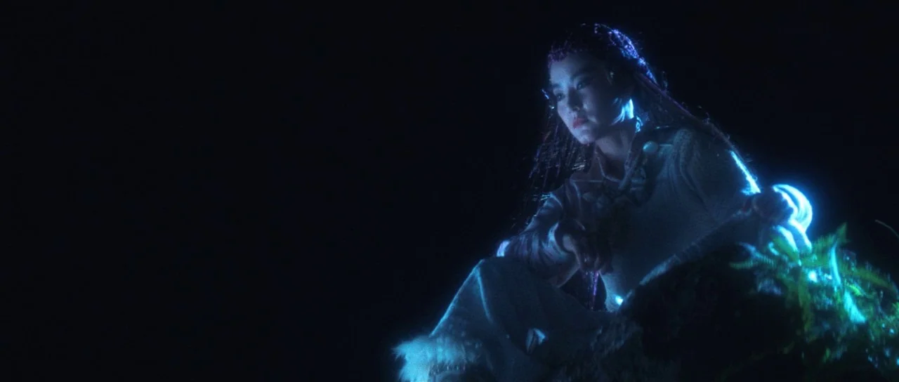 The Bride With White Hair (1993) movie still: medium — A woman sits in a dark, outdoor setting with ferns partially obscuring the foreground. She…