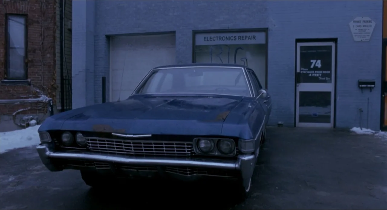 Foolproof (2003) movie still: wide — A dark blue vintage sedan sits in a paved lot before a brick building. The building facade features a window…
