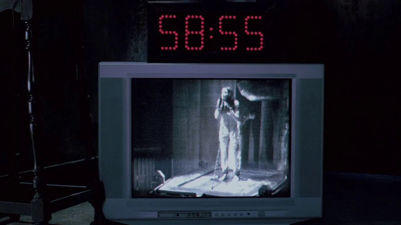 Saw VII: The Final Chapter (2010) movie still: closeup — A dark room contains an old television monitor showing black-and-white video footage. A…