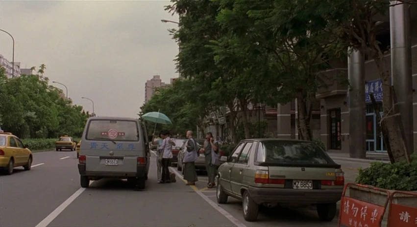 Yi-Yi (A One and A Two) (2000) movie still: wide — A suburban street lined with mature trees and parked cars under an overcast sky. Two monks and a…