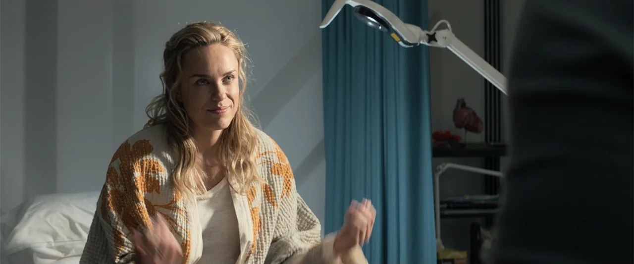 The Meg (2018) movie still: medium — An exam room with a teal curtain and a professional medical lamp in background. A blonde woman in a patterned…