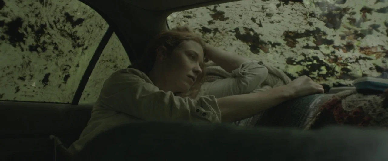 Here Alone movie still: medium — The interior of a car features mud-splattered windows obscuring the view outside. A woman rests her head on her…