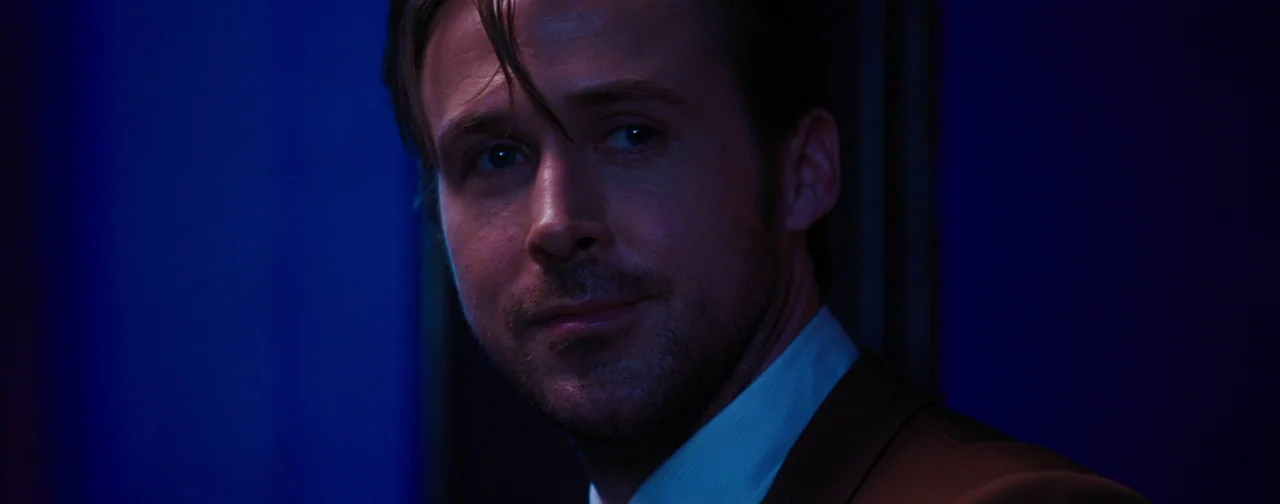 La La Land (2016) movie still: closeup — A dimly lit interior space is illuminated by intense blue and purple light sources. A man in a brown suit…