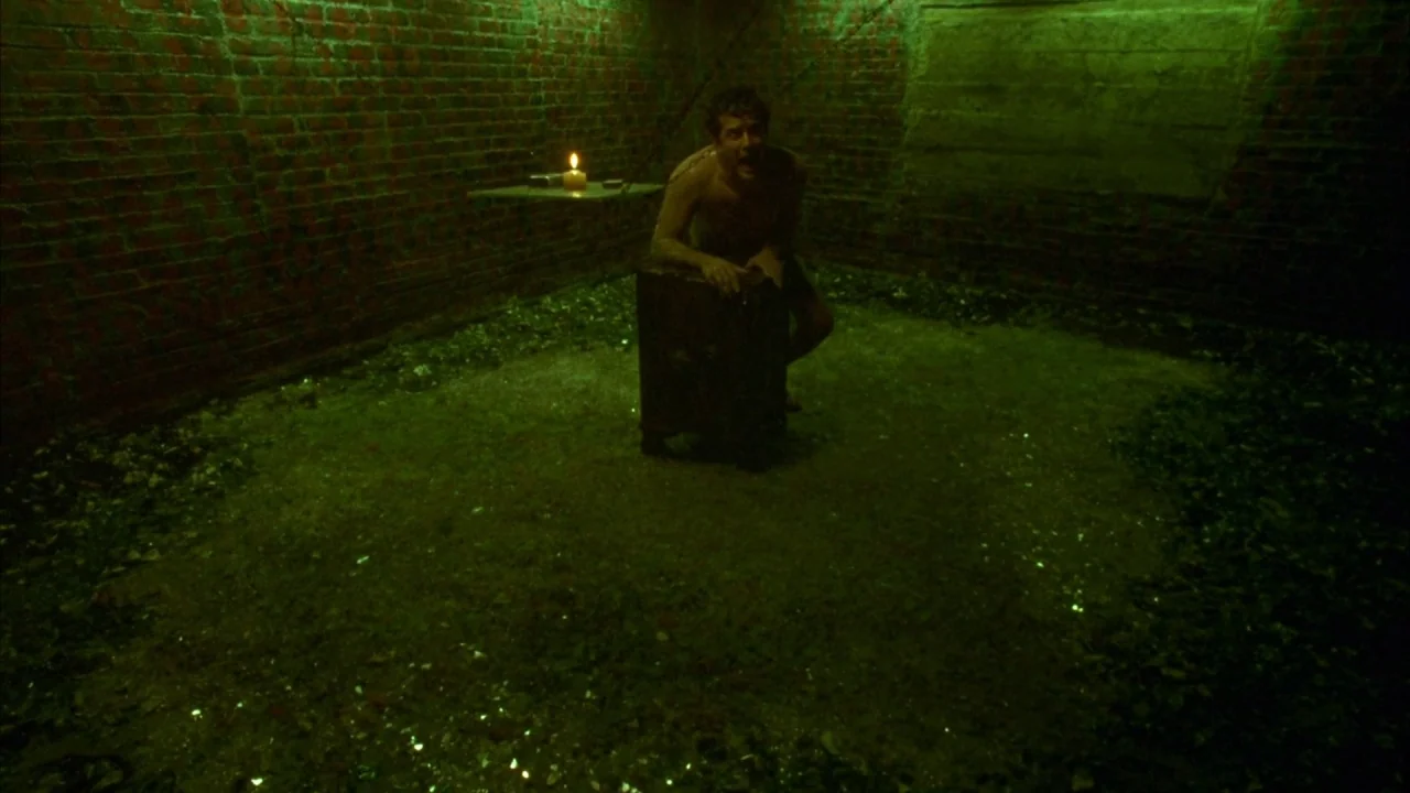 Saw movie still: fullbody — A dark subterranean space features rough brick walls and a debris-covered concrete floor. A man kneels in the center of…