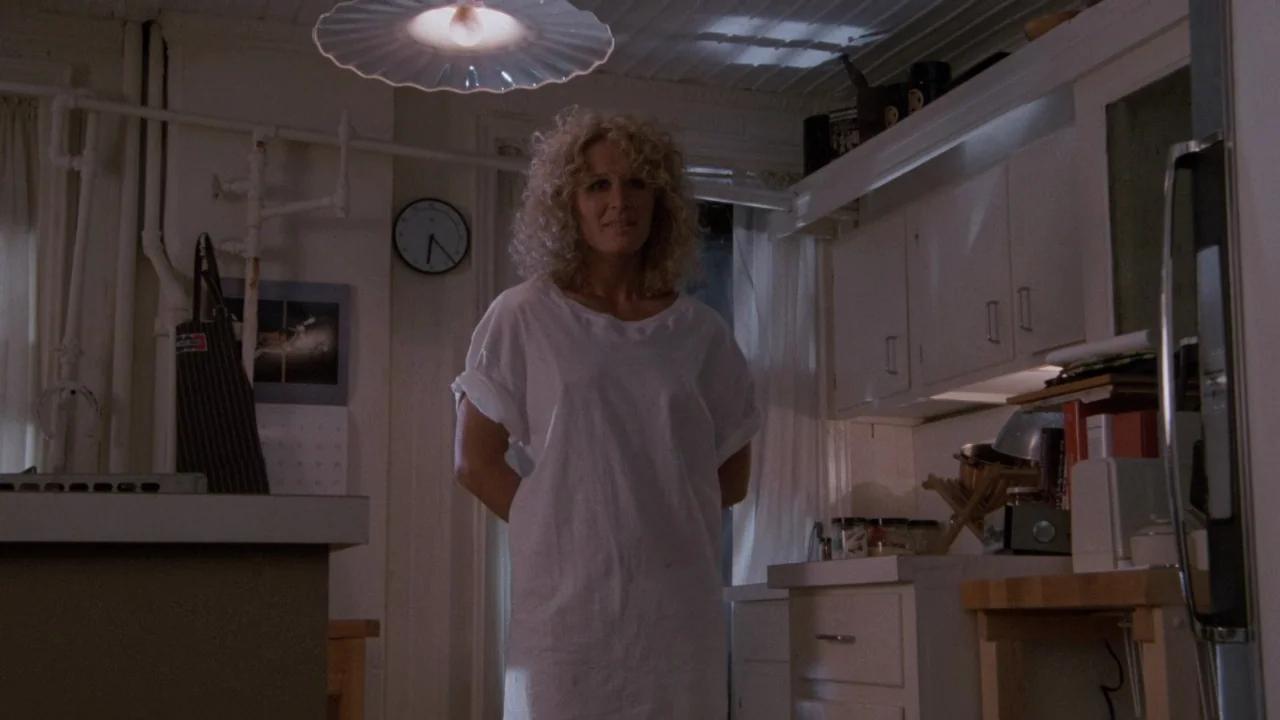 Fatal Attraction (1987) movie still: medium — A dimly lit room with off-white cabinets and a simple wall clock. A blonde woman in a white nightshirt…