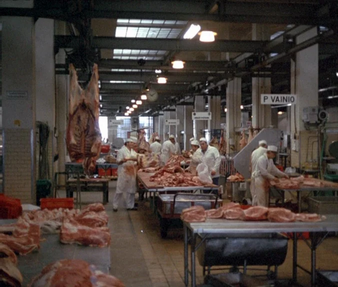 Crime & Punishment (1983) movie still: wide — A spacious industrial processing floor contains multiple stainless steel tables covered in raw meat…