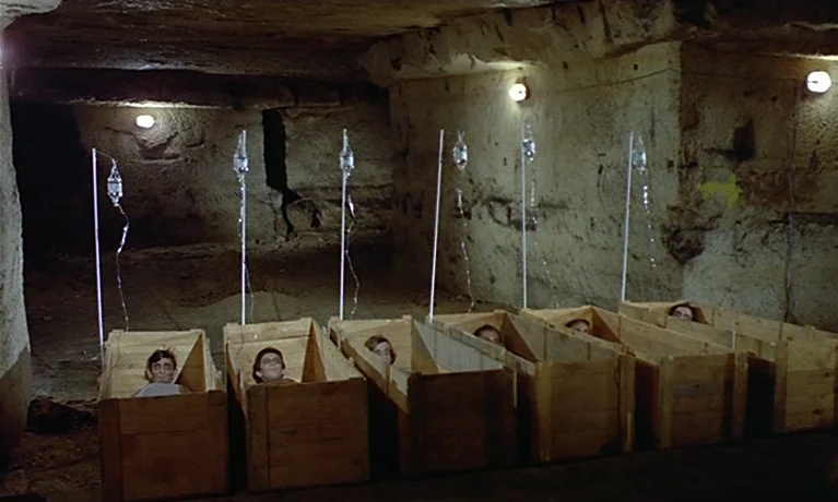 Nuits Rouges (1974) movie still: wide — A dark subterranean tunnel with rough stone walls and low ceilings features multiple wooden crates. Several…