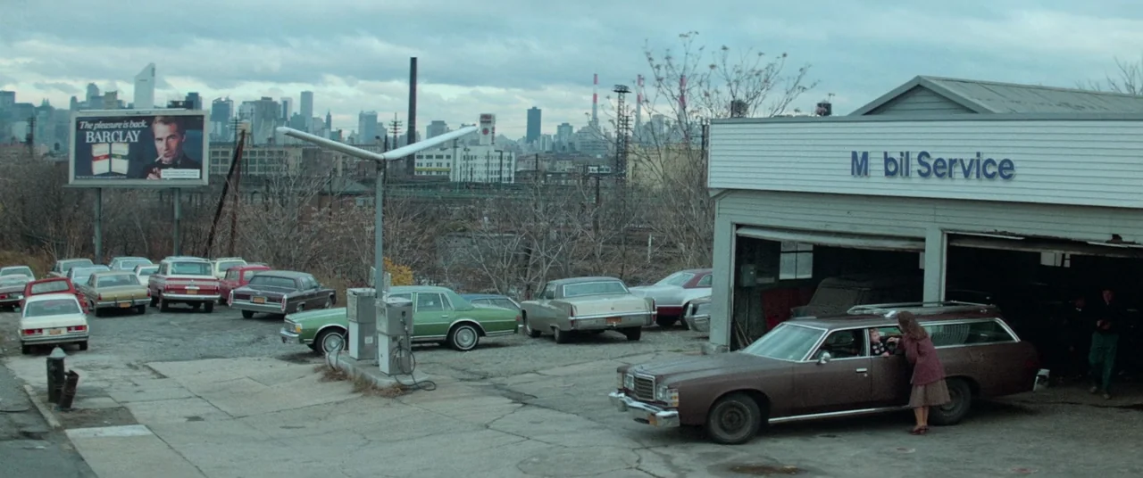 Vigilante (1982) movie still: wide — An outdoor gas station lot under an overcast sky with a distant city skyline. A woman talks to someone inside a…