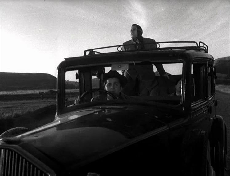 I Vitelloni (1953) movie still: medium — A dark vintage car travels along a desolate road under a clear sky. Three men are visible inside, one…