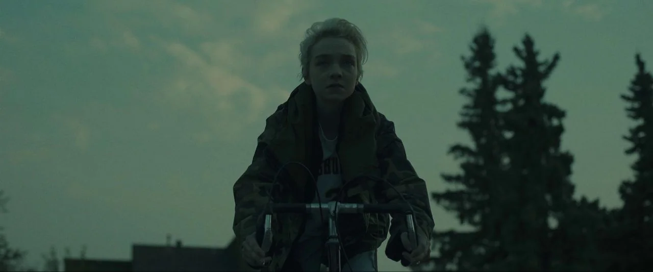 Come True (2020) movie still: medium — An outdoor suburban road at dusk with evergreen trees silhouetted against a fading sky. A young person with…