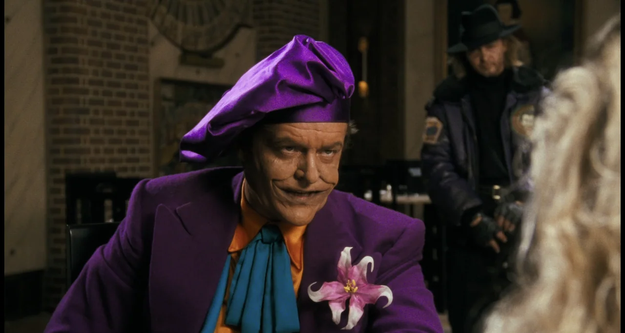 Batman (1989) movie still: medium — A dimly lit room features a man in a vibrant purple suit seated at dinner. He sits across from a companion while…
