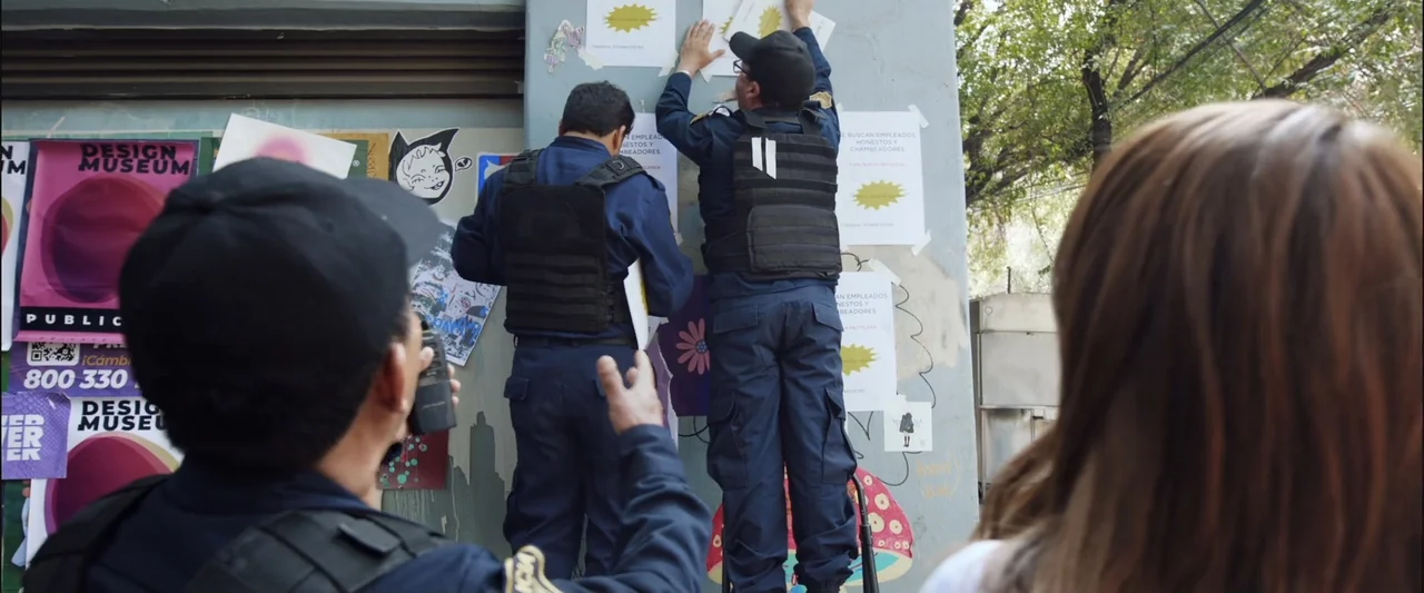 Winner Takes the Cake (2025) movie still: medium — A group of people stand before a weathered urban concrete wall covered in posters. Two uniformed…