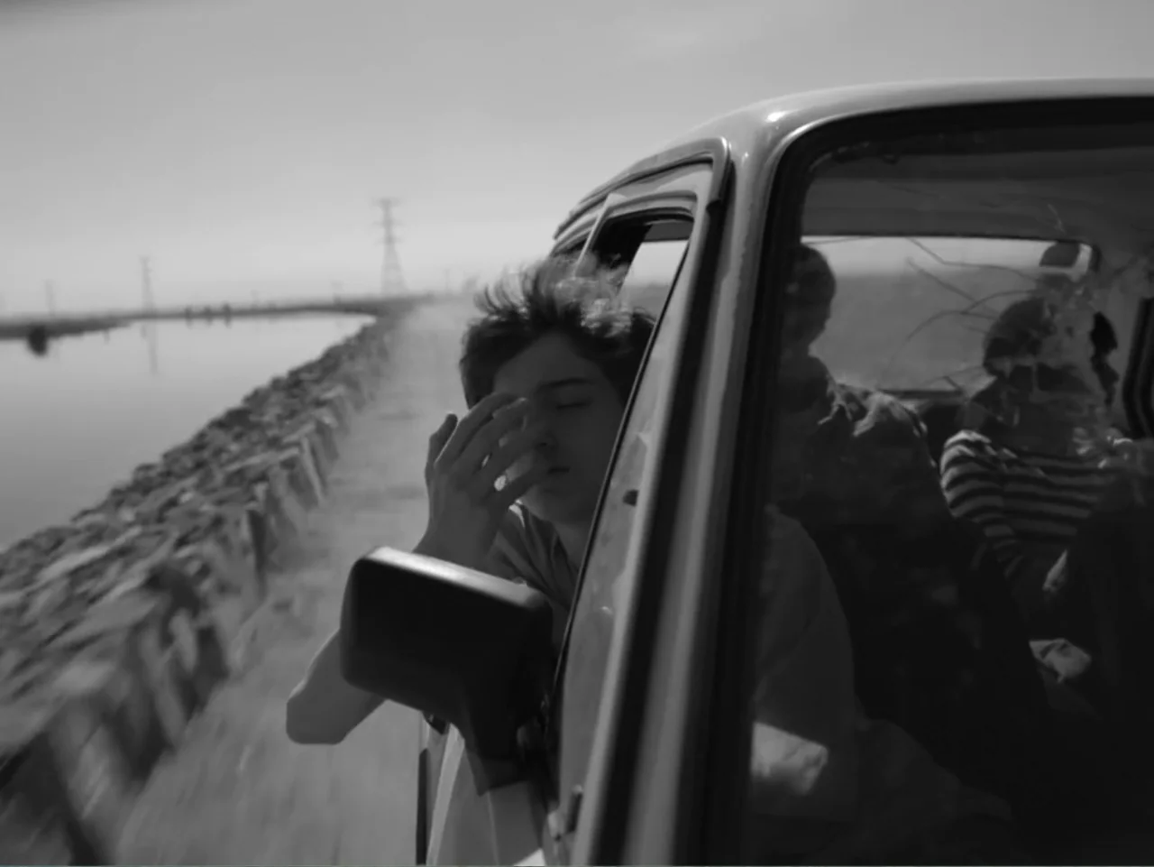Güeros (2014) movie still: medium — A moving car drives alongside a water channel with utility towers in the distance. A young man rests his head…