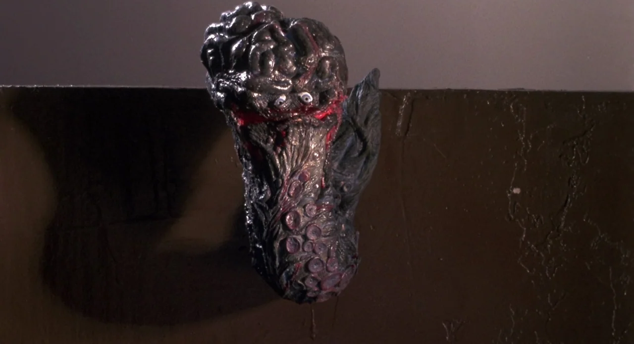 Brain Damage (1988) movie still: closeup — A dark, textured organic creature with a brain-like top sits against a wall. The creature has multiple…