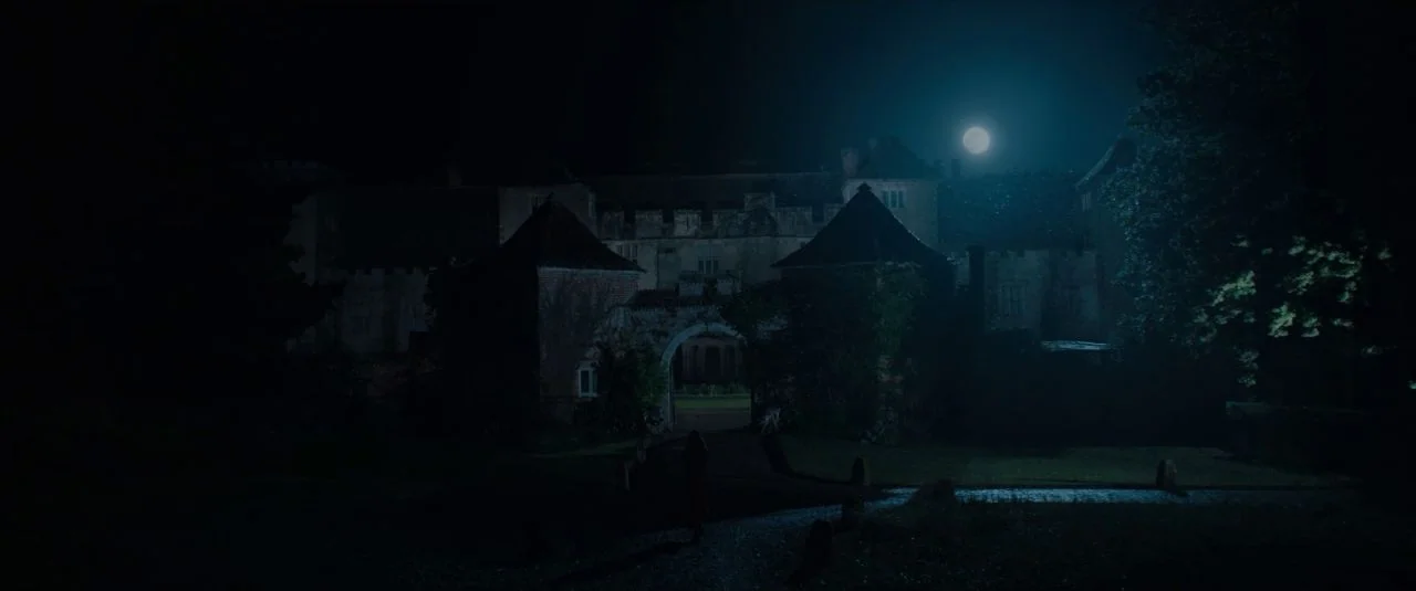 Rebecca movie still: establishing — An ancient stone manor sits under a dark night sky illuminated by a moon. A large arched entryway leads into the…